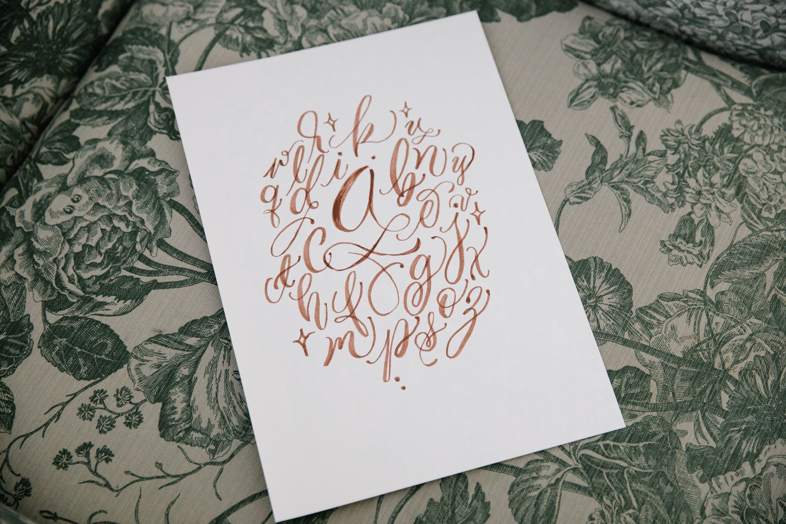 A beautiful handwritten alphabet on paper placed on a floral textile background, showcasing artistic calligraphy.